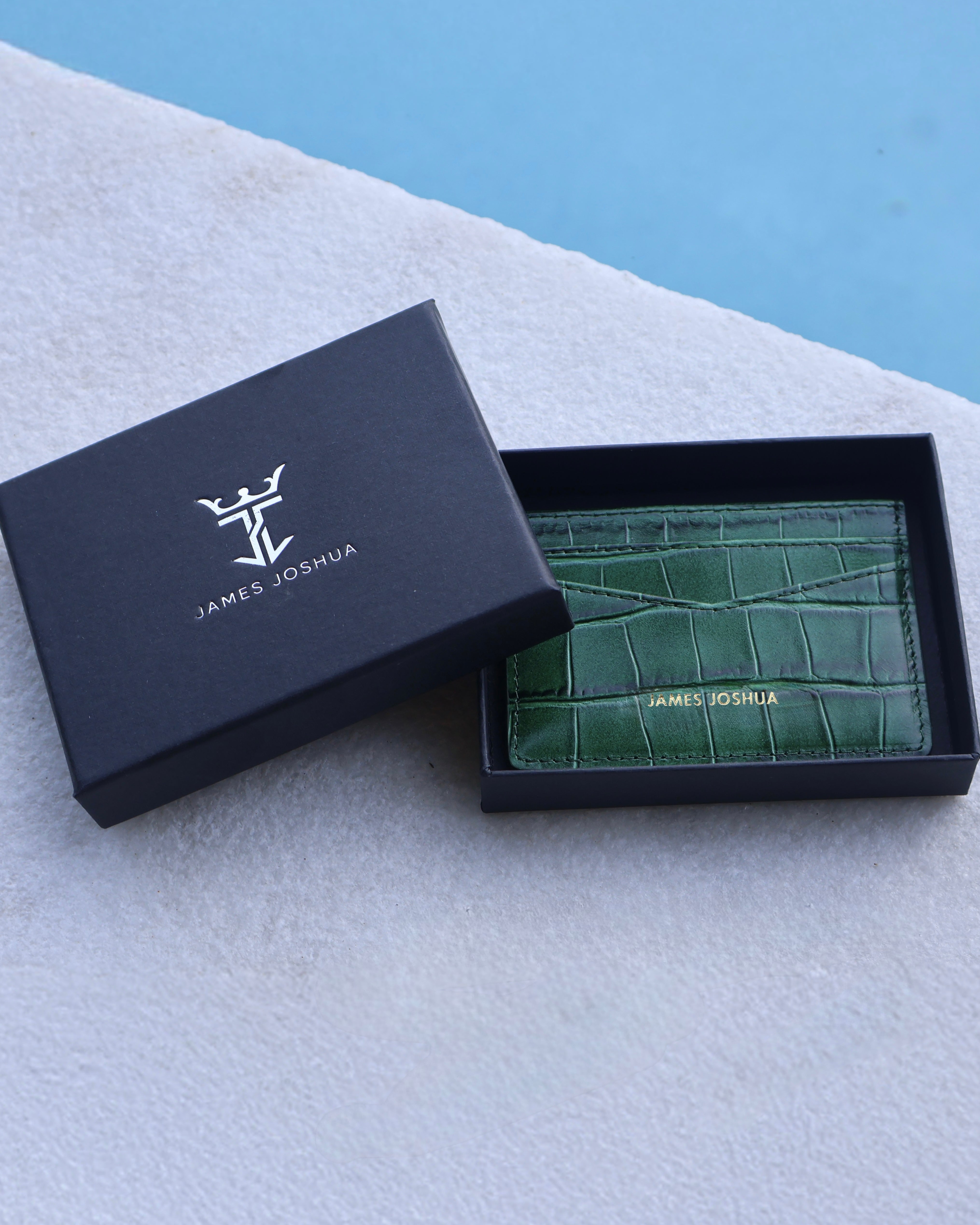 Green crocodile-textured cardholder in a black 'James Joshua' box on a light gray surface with a blue background.