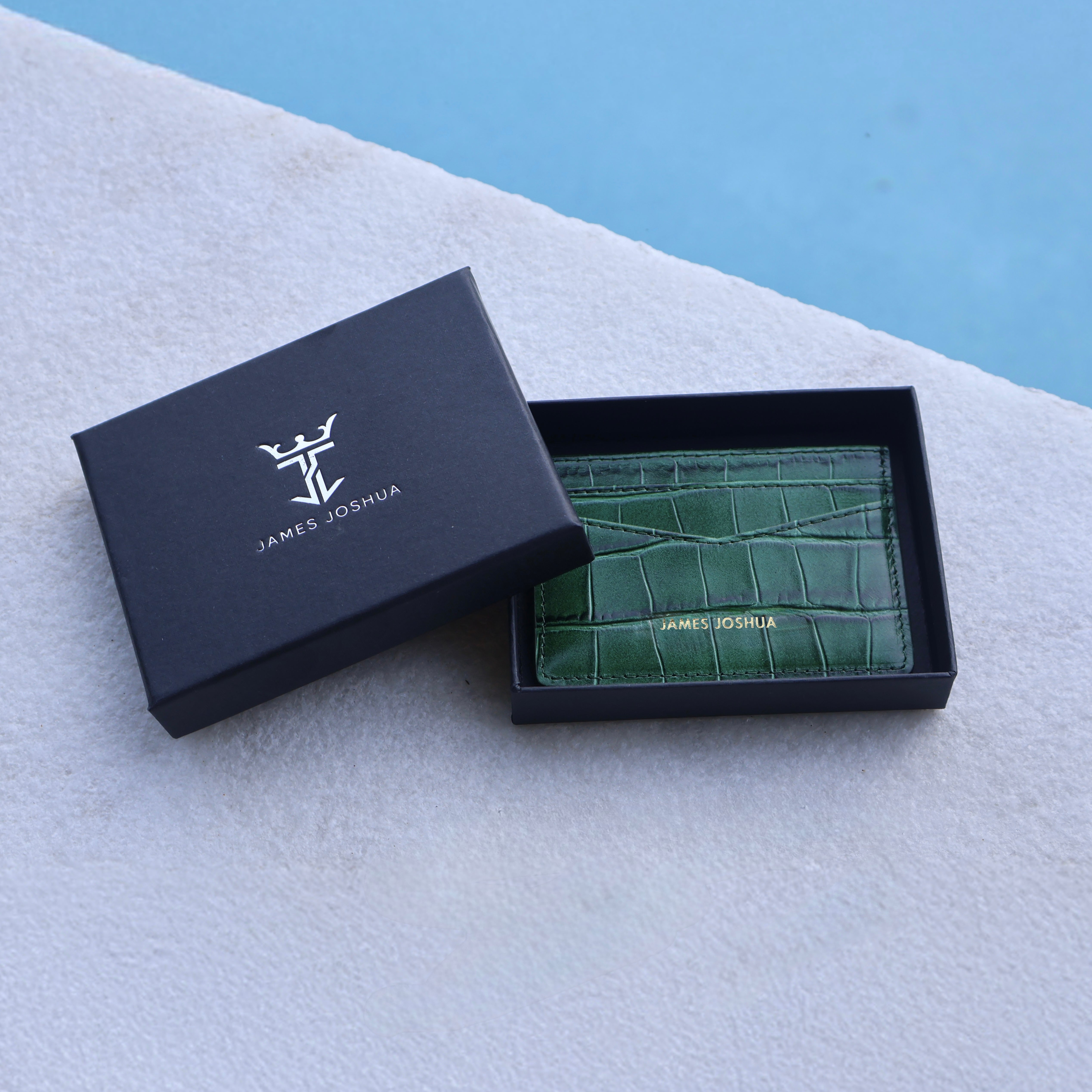 Green crocodile-textured cardholder in a black 'James Joshua' box on a light gray surface with a blue background.